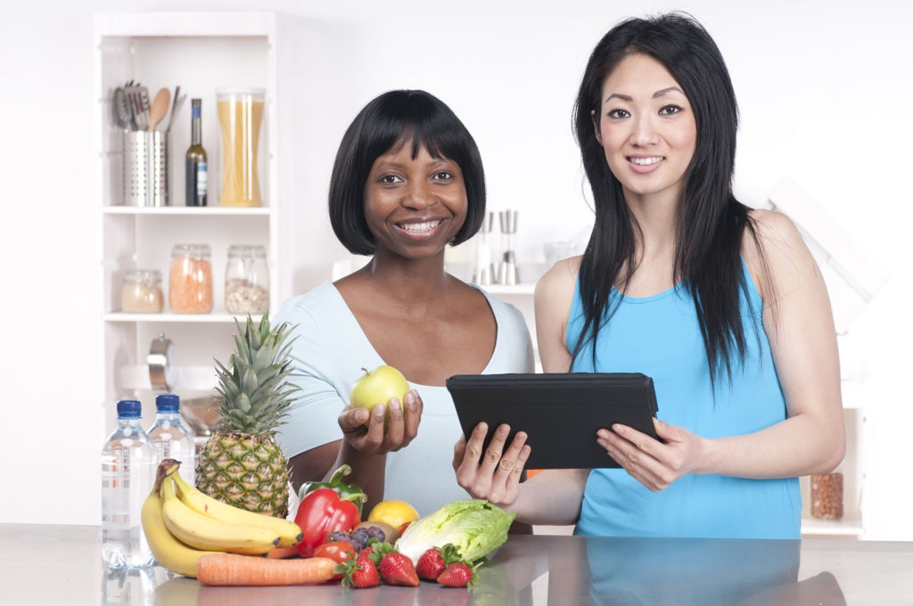 Two women in a kitchen with fruits and vegetables on a table whilst looking up information on a computer tablet.See more healthy diet images here: